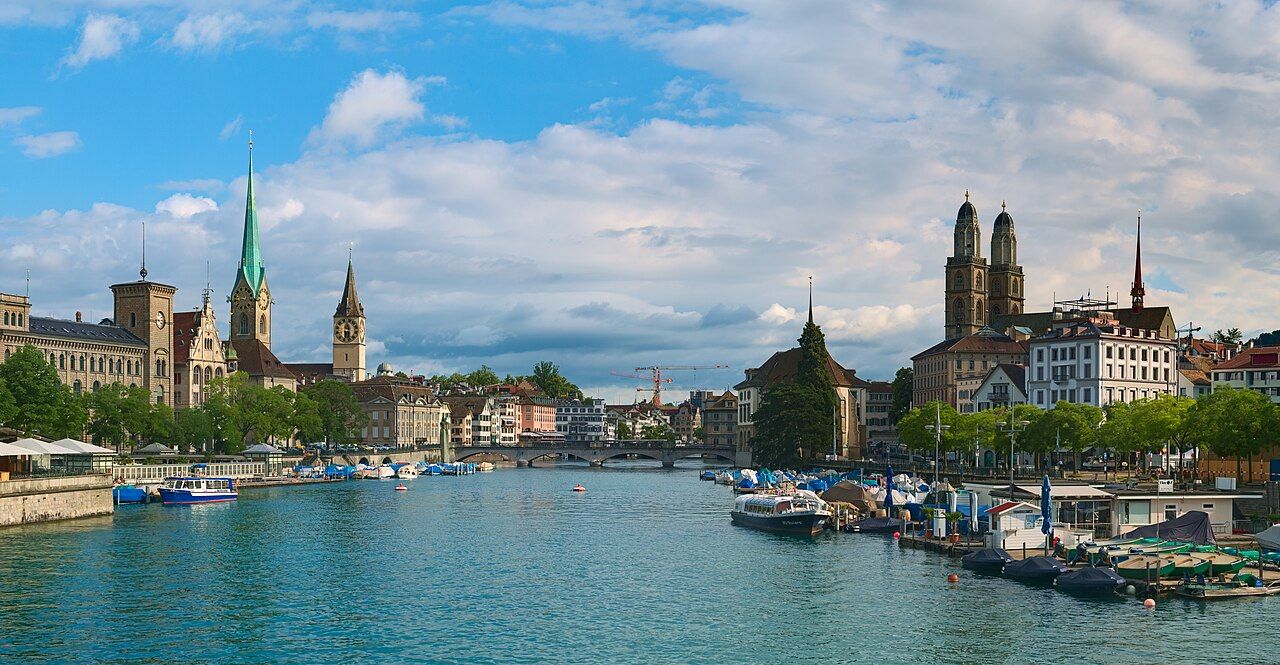 Pohled na Curych z mostu Quaibrücke: věže kostelů, řeka Limmat a v pozadí jezero s horami
