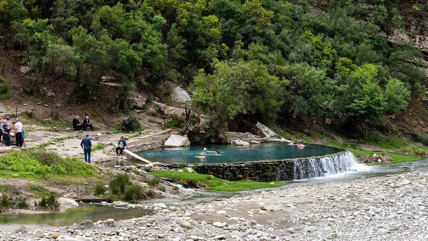 Termální prameny Bënjë u Përmetu — kamenná káď s horkou vodou a koupající se lidé